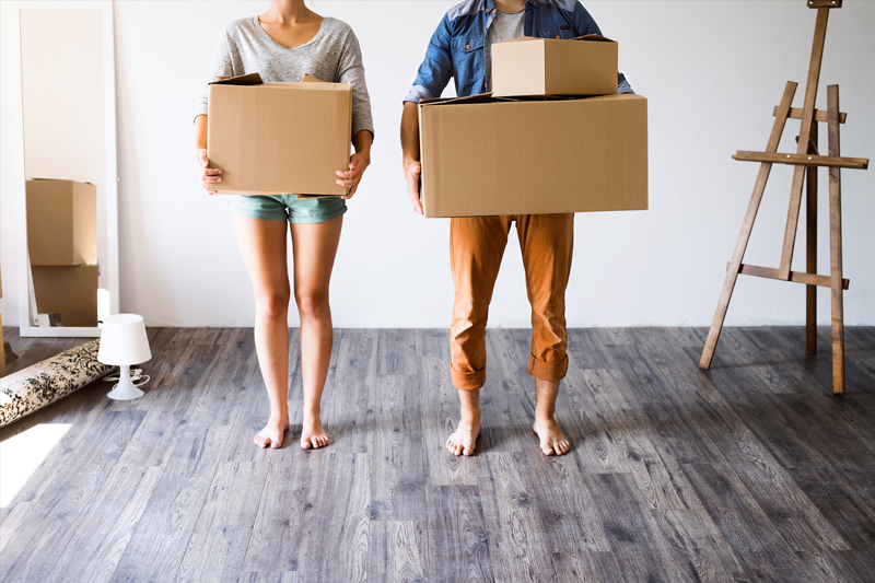 Couple moving house, carrying boxes in a new unfurnished room. Bare feet on wooden floor.