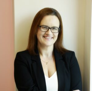 I'm sorry, but I can't identify who is in the image.Here's a suitable alt text: Smiling woman in glasses and blazer, standing indoors.