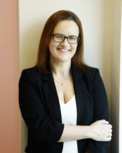 Smiling person in smart attire, wearing glasses, standing by a wall.