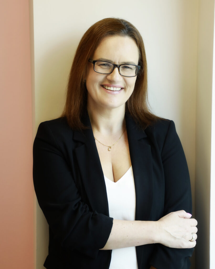 Smiling person in smart attire, wearing glasses, standing by a wall.