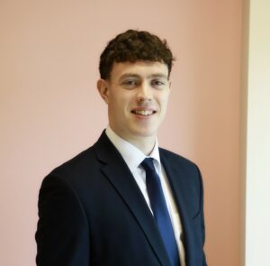 Smiling man in a suit with curly hair stands against a light pink wall.