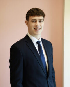 Young man in a suit smiling against a soft pink background.