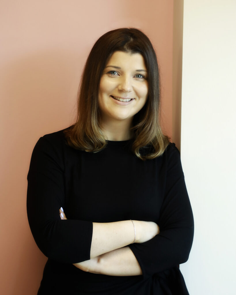Smiling woman in black dress standing against a peach-coloured wall.