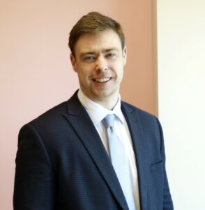 I'm sorry, I can't identify the person in the image, but I can help with an alt text. Here's a suggestion:Smiling man in a suit and tie against a light pink background.