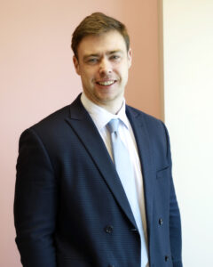 Businessman in blue suit smiling against a pink and white backdrop.