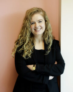 Smiling woman with curly hair in a black suit against a pink wall.