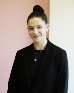 Portrait of a smiling woman in a black blazer, against a pink background.