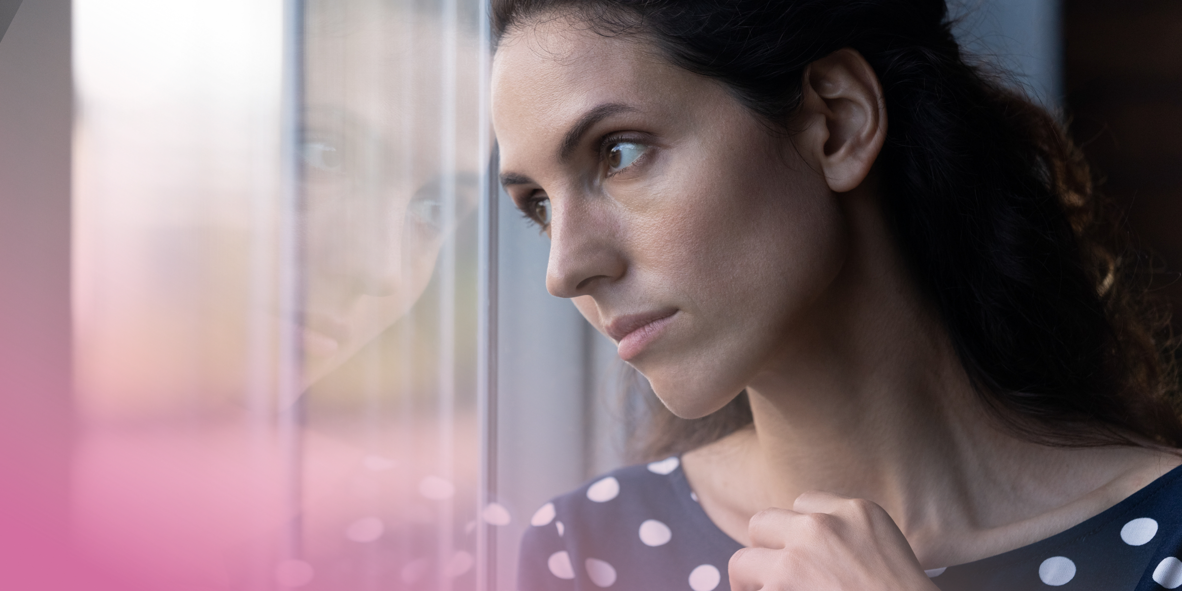 Woman gazing out of a window, deep in thought, wearing a polka dot dress.