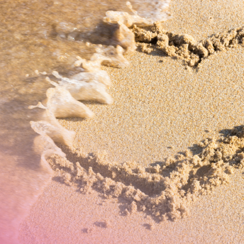 Heart shape drawn in sand as a wave washes over, symbolising fleeting moments by the seaside.